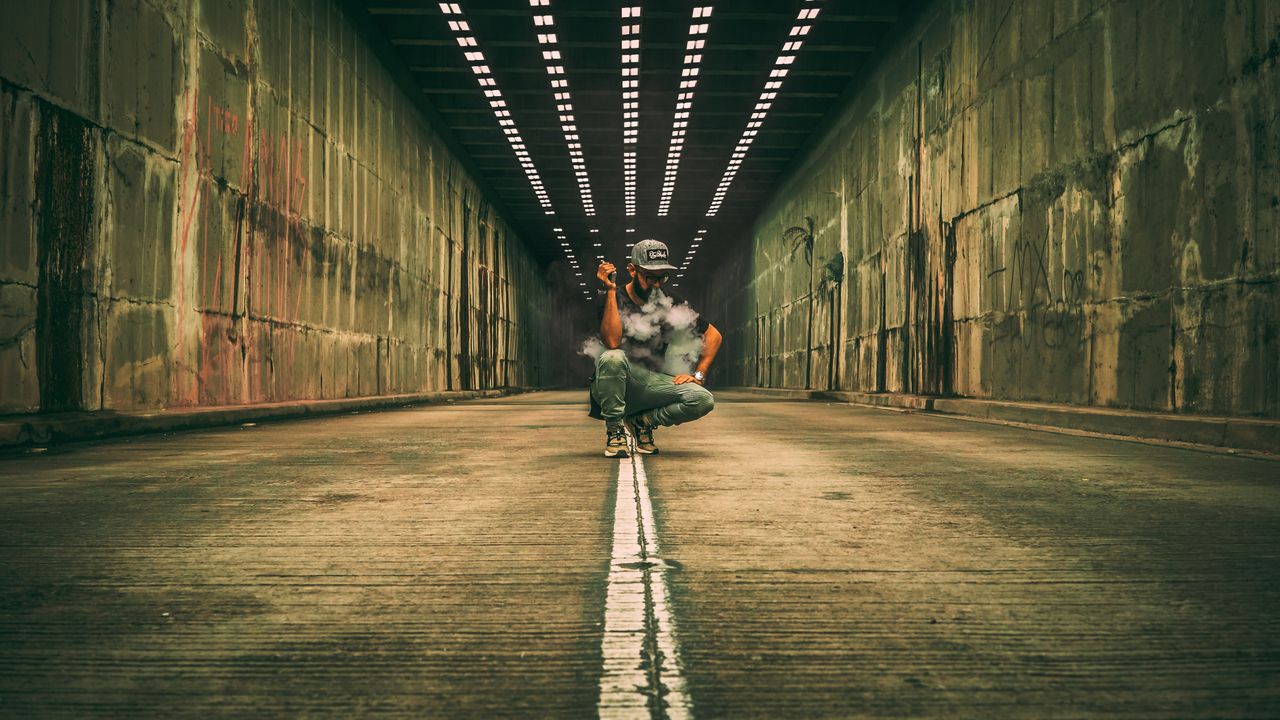 man squats in a tunnel vaping surrounded by smoke and moody lighting