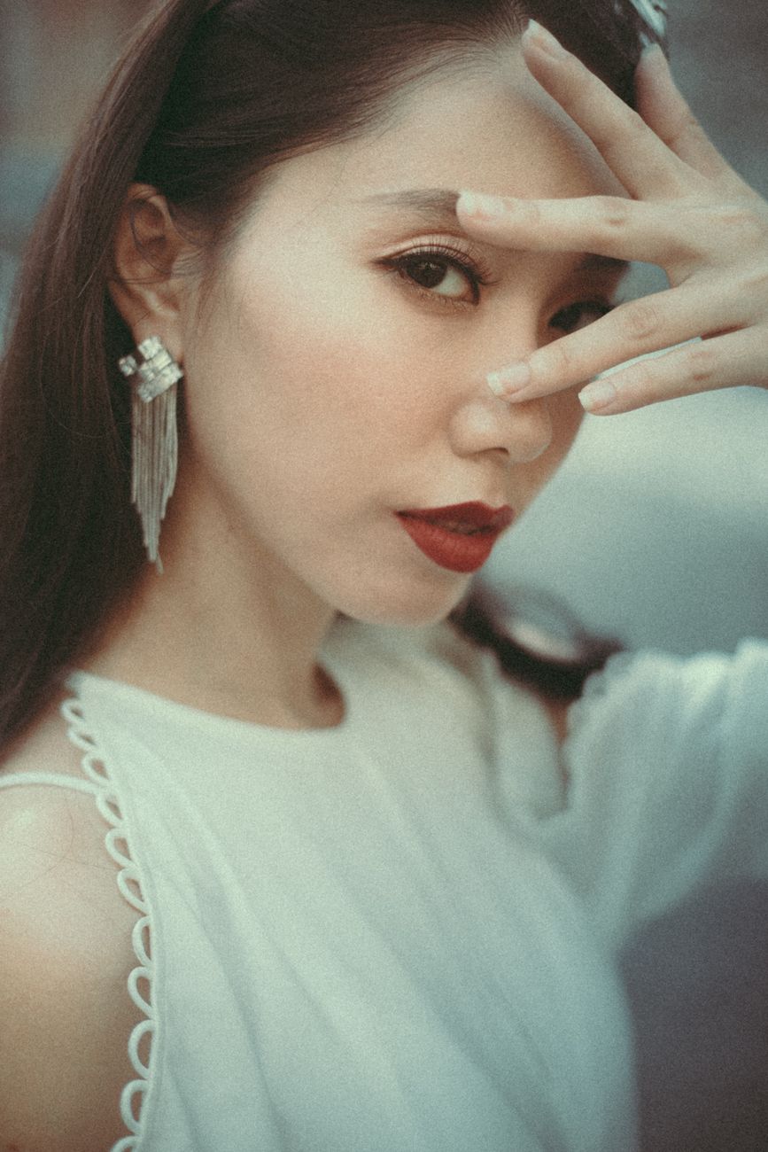 Crop modish Asian female in trendy white dress and earrings looking at camera through spread fingers