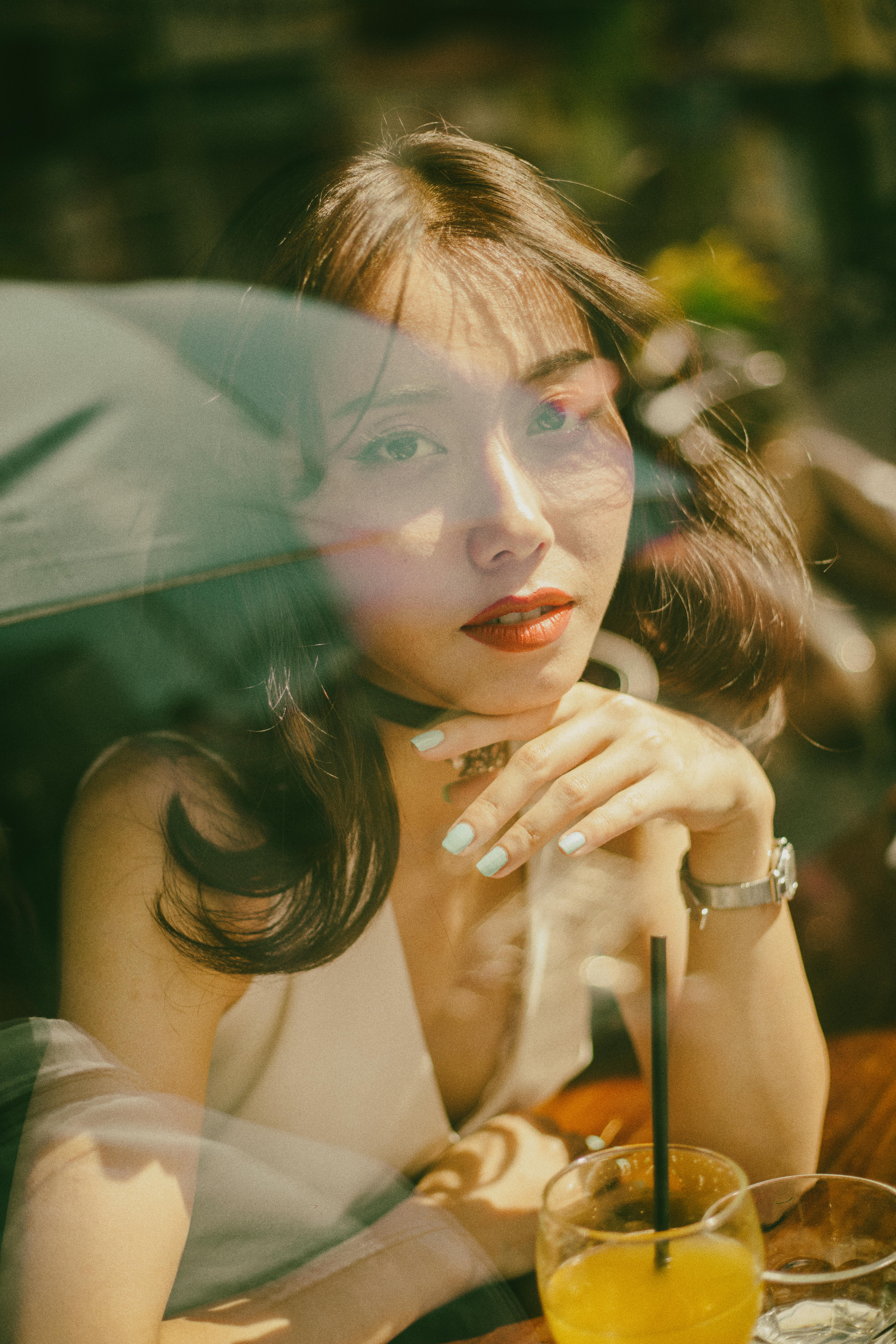 Through glass of pretty Asian female sitting at table in cafeteria with glass of cocktail and touching chin tenderly while looking at camera dreamily