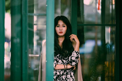 Calm pondering female in stylish dress standing near glass doorway with hand raised and looking at camera