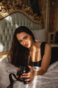 Elegant woman lying on bed holding a camera in a warmly lit bedroom.