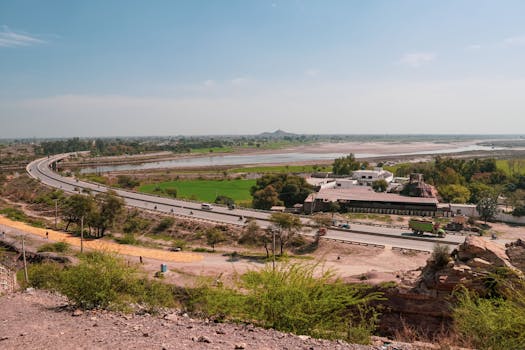 Wide-angle view of highway curving through scenic landscape with river and lush fields under a clear blue sky.