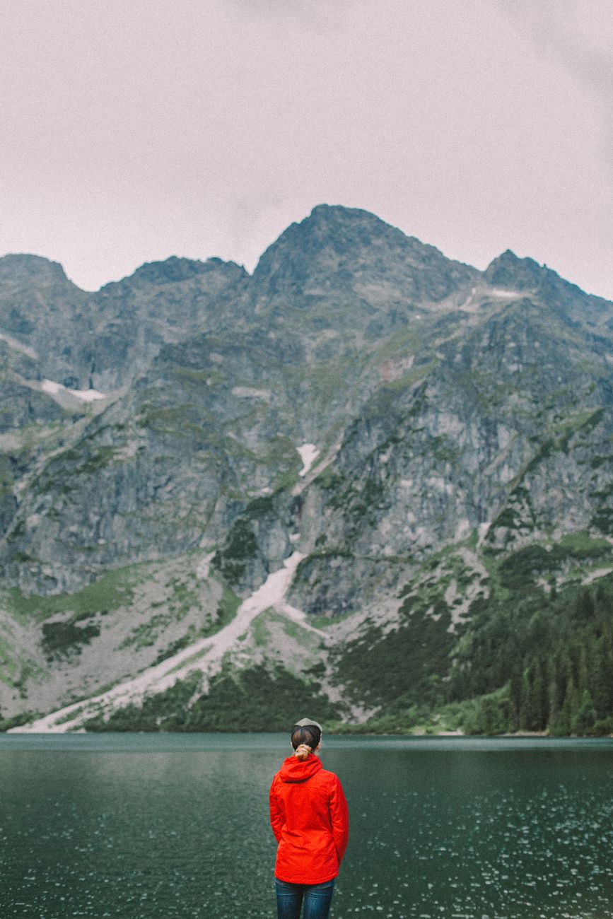 Traveler in a red jacket gazing at a peaceful mountain lake surrounded by lush green mountains.