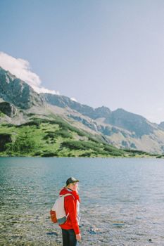 在清澈的蓝天下，一位穿着红色外套、戴着帽子的旅行者正探索宁静的山间湖泊