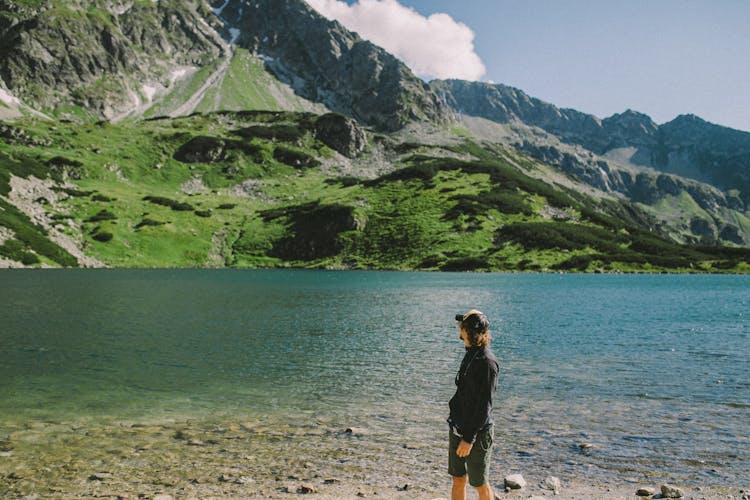 A Man In Black Long Sleeves Standing Near The Lake While Looking At The Mountain