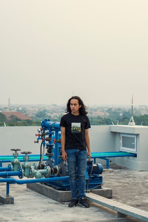 Free A young man with long hair stands on a rooftop with a city view, next to industrial pipes. Stock Photo