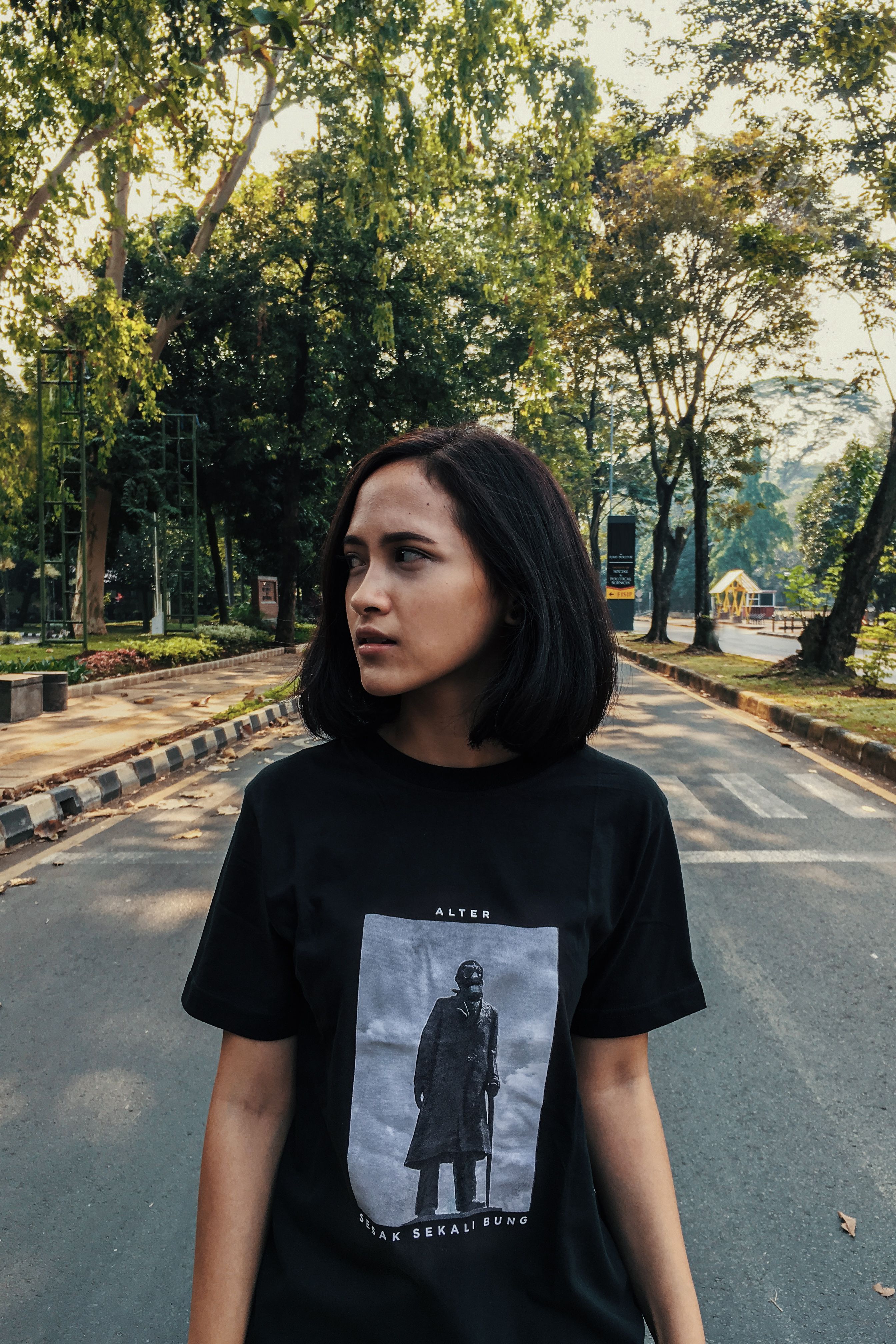 Portrait of a young woman in a black shirt standing on a street in Indonesia surrounded by trees.