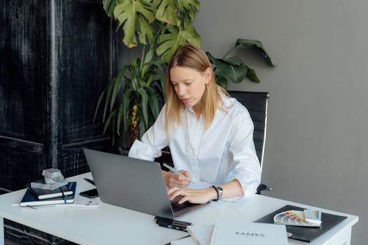 Focused woman in white shirt working on laptop at desk with plants and books.
