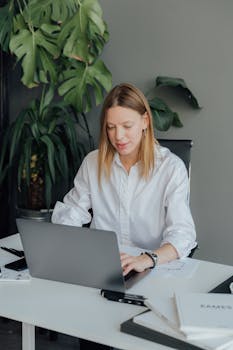 Focused young woman working on a laptop in a stylish office environment with greenery.
