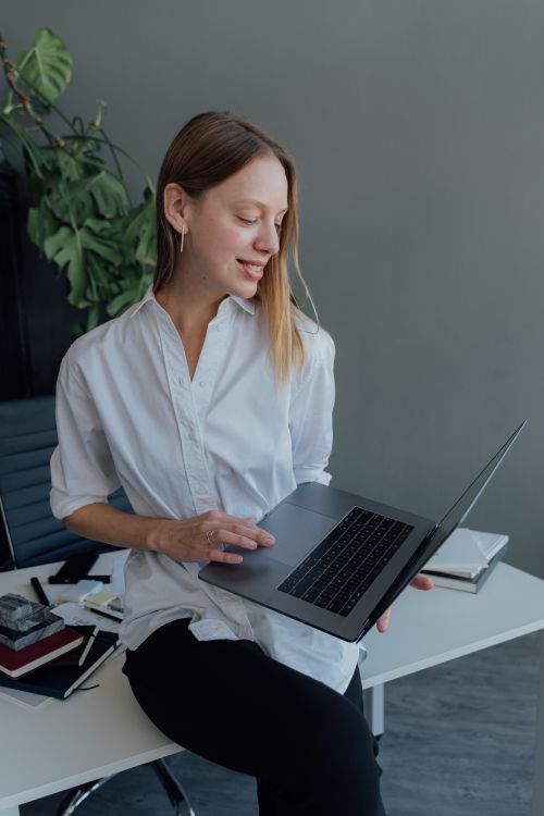 Free Woman in white shirt using laptop at home office desk, surrounded by notebooks and digital devices. Stock Photo