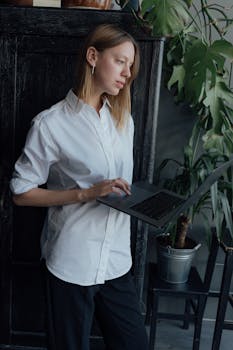 Woman in white shirt using laptop near potted plant indoors, illustrating remote work.