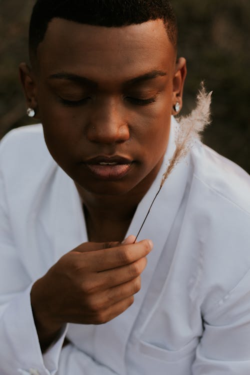 Dreamy young African American male in white jacket with short hair and small glistening earrings thoughtfully looking at delicate beige fluffy feather on blurred background