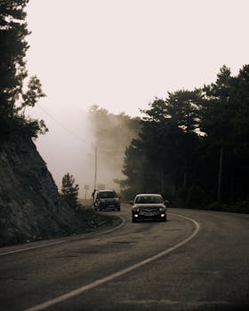 A misty mountain road with cars driving through a dense forest area.