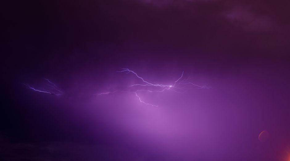 A vivid purple sky illuminated by a striking lightning bolt during a thunderstorm at night.