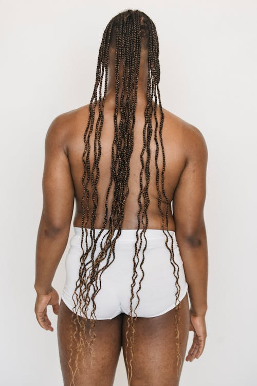 Back view of unrecognizable plump African American male with Afro braids wearing white underpants standing on white background in light studio