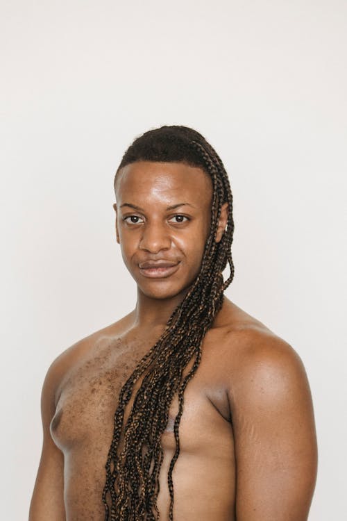 Content feminine African American male with naked torso and Afro braids looking at camera while standing on white background in studio