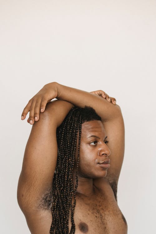 Side view of African American male with Afro braids and naked torso keeping crossed arms on head while standing against white background