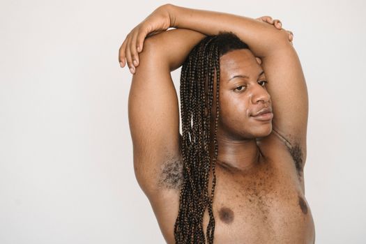 Feminine African American male with naked torso standing on white background with crossed arms on head and hairy armpits in studio