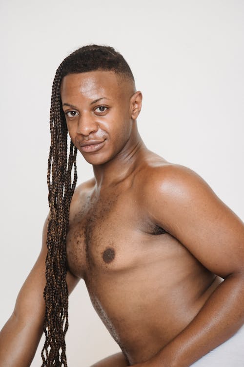 Optimistic African American male with Afro braids and naked torso looking at camera while sitting on white background with arm bent in elbow