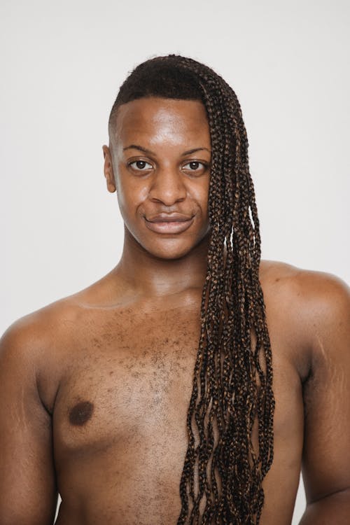 Confident androgynous feminine African American male with naked torso looking at camera while standing on white background in light studio