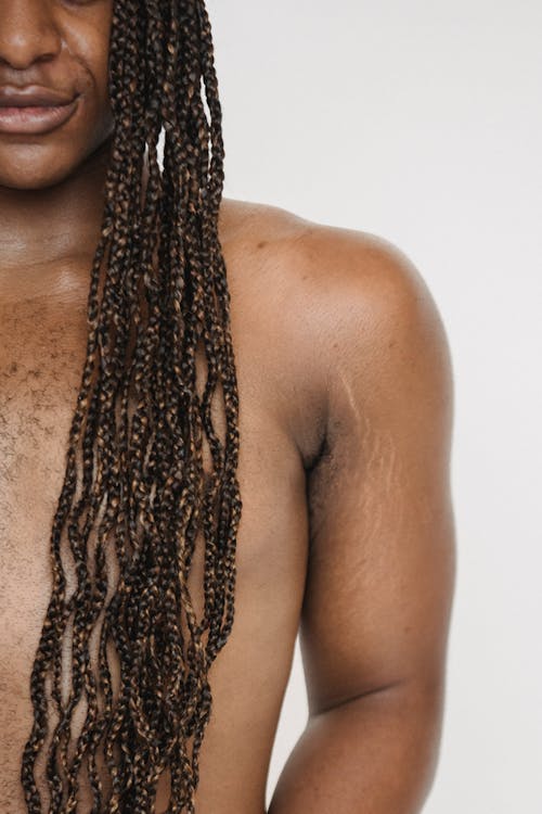 Unrecognizable queer androgynous African American male with naked torso and long Afro braids standing on white background in modern studio