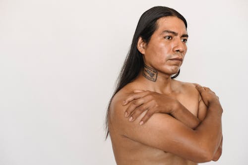 Emotionless adult American Indian male with long hair crossing arms on naked torso and looking away while standing against white wall in studio