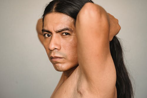 Side view of crop young Native American male with hands behind head looking at camera on light background