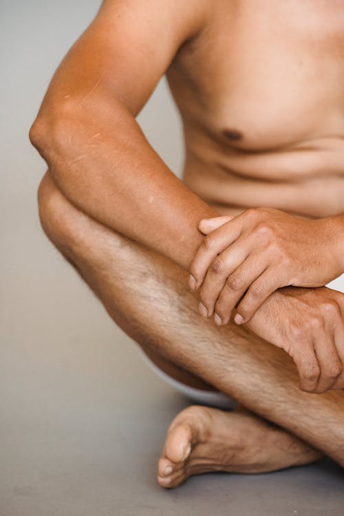 Crop anonymous barefoot male wearing white underpants sitting on floor with legs crossed against light wall in studio