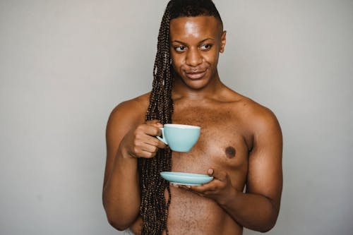 Free Crop African American male with long braids standing with naked torso and drinking from ceramic cup in studio while looking away with pouted lips Stock Photo