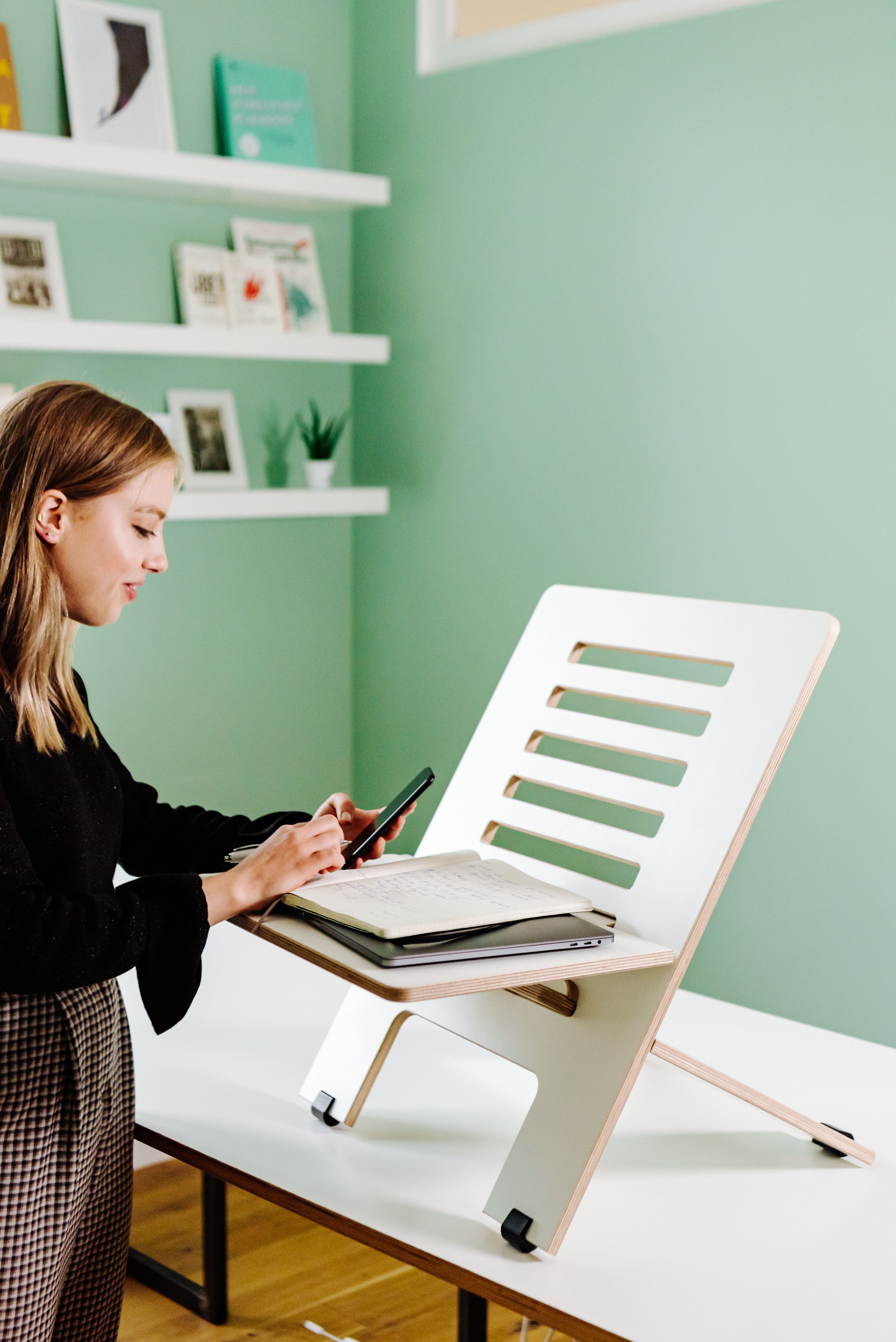 Young woman using a phone at home office with adjustable desk stand and notebooks. - How to Set Up an Ergonomic Home Workspace on a Tight Budget