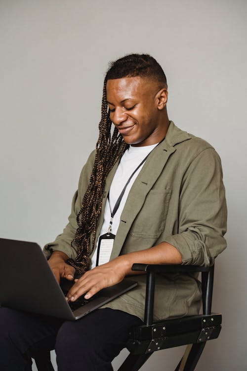 Positive African American male with Afro braids and name tag typing on computer while sitting on white background in studio