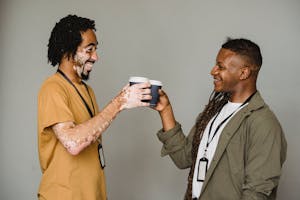 Side view of cheerful androgynous man with Afro braids standing on gray background near black coworker with vitiligo skin during coffee break