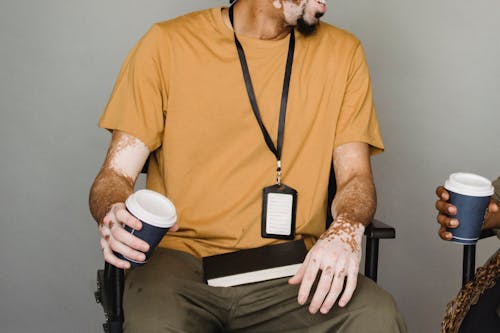 Free Unrecognizable male with skin disease wearing name tag sitting on gray background with cup of takeaway beverage and notebook near ethnic anonymous coworker during coffee break Stock Photo