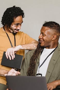 Two professionals with laptops share a cheerful moment in an office environment.