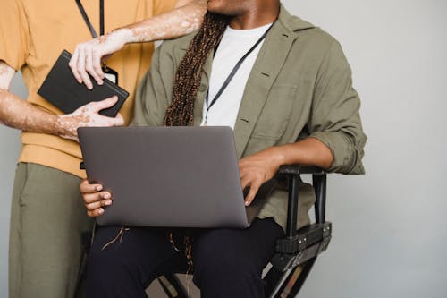 Unrecognizable African American male with Afro braids typing on computer while sitting on gray background near colleague with vitiligo and notepad