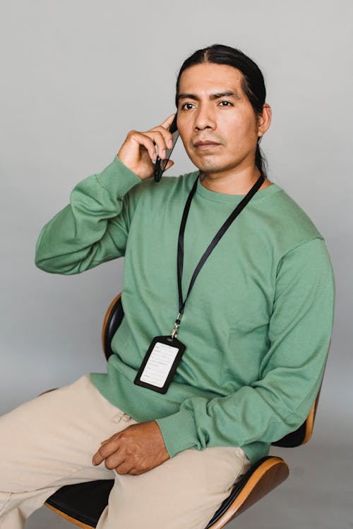 Confident Indian male wearing name tag and speaking on smartphone while looking away and sitting on gray background in studio during work