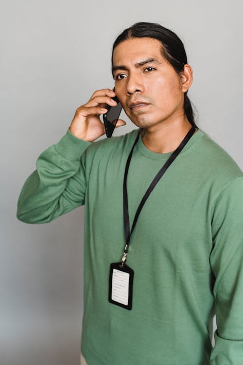 Concentrated Indian male wearing id card standing on gray background and looking away while having phone conversation on smartphone in studio