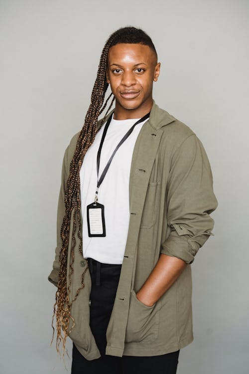 Androgynous African American male with Afro braids wearing id card looking at camera while standing on white background with hand in pocket