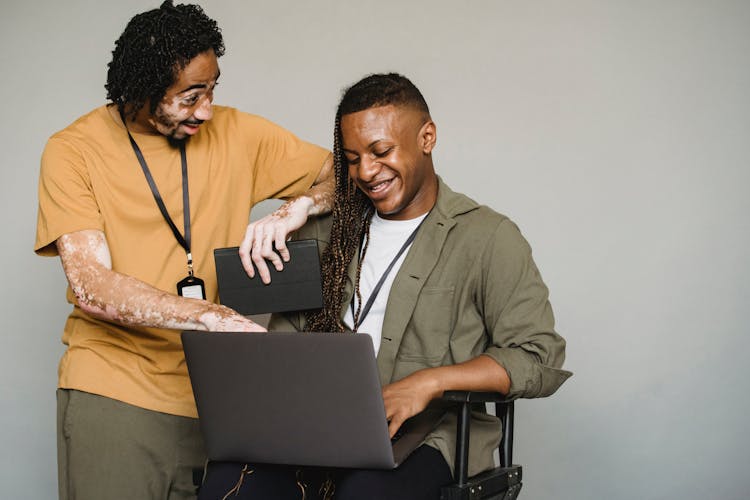 Cheerful Man With Vitiligo Looking At Androgynous Black Coworker With Computer