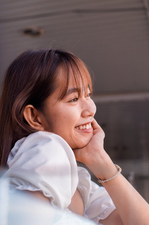 Side view of dreamy Asian female in white apparel leaning on hand and looking into distance while standing near blurred railing