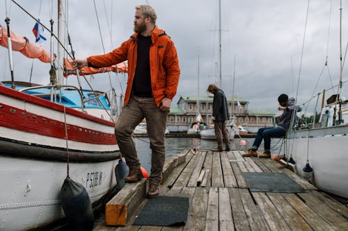 бесплатная Мужчина в оранжевом худи и черных штанах стоит на деревянной пристани Стоковое фото
