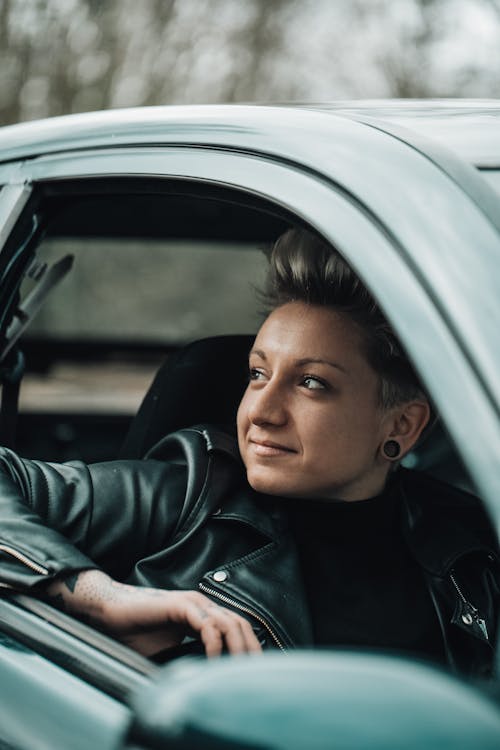 Attractive young female with trendy haircut in black leather jacket sitting in automobile and smiling while looking away