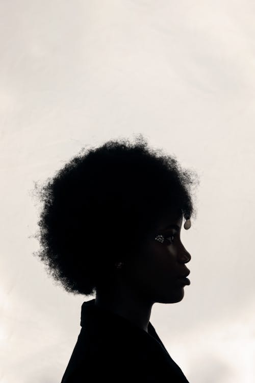 Side view of calm young black woman with Afro hair and creative accessories standing against gray sky and looking away pensively