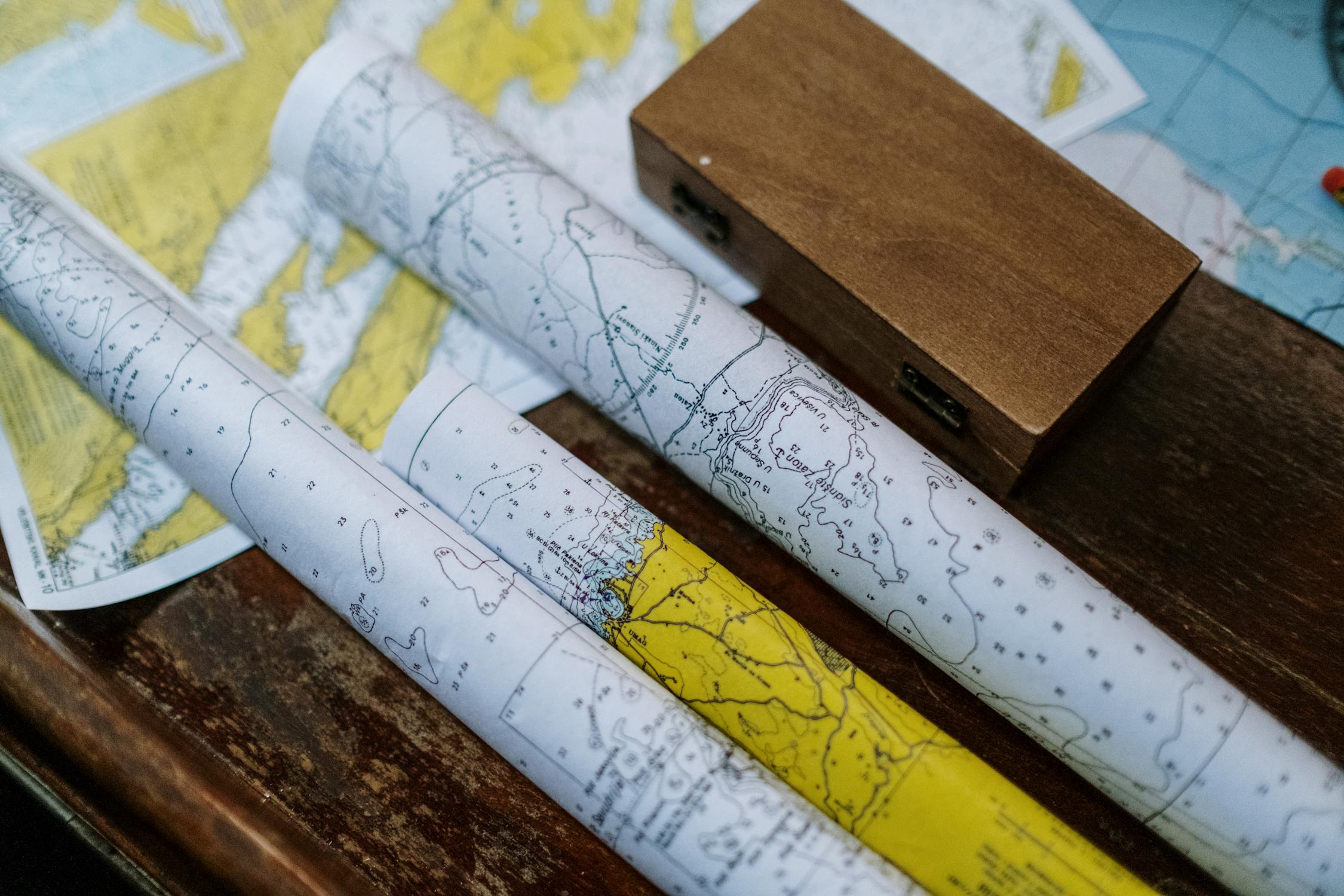 Close-up of rolled nautical maps on a wooden surface, with a small wooden box nearby and a partially visible world map underneath.