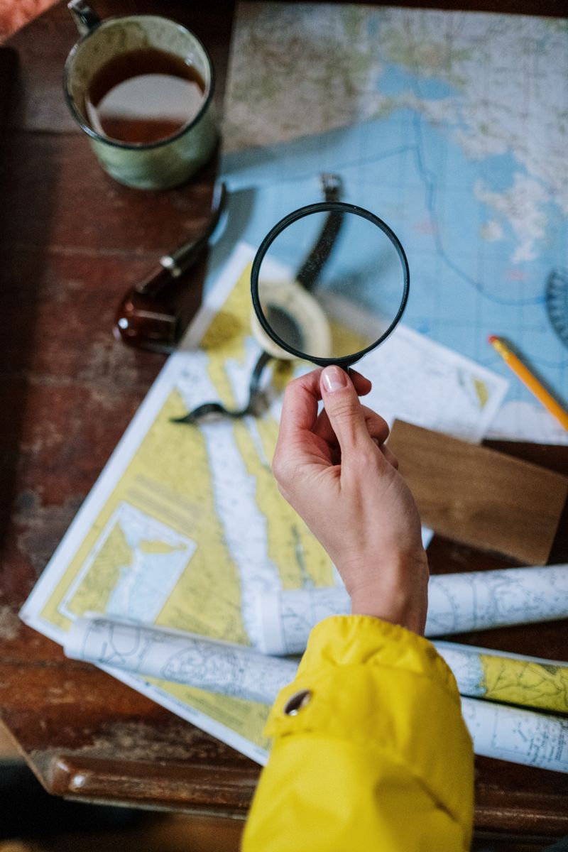 A person's hand with a magnifying glass examining nautical maps on a wooden table.