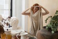 Young female artisan with closed eyes putting on apron near table with jug and glassware in workshop