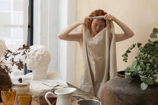 Young female artisan with closed eyes putting on apron near table with jug and glassware in workshop