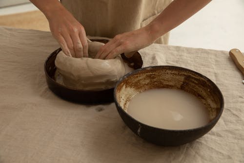 From above of crop anonymous artisan sculpting soft clay near bowl with liquid on table in workroom