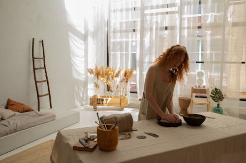 Concentrated redhead female wearing light apron kneading clay in baking pan while creating in modern apartment