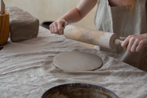 Crop anonymous female artisan in apron rolling out clay slab on table while creating in light pottery